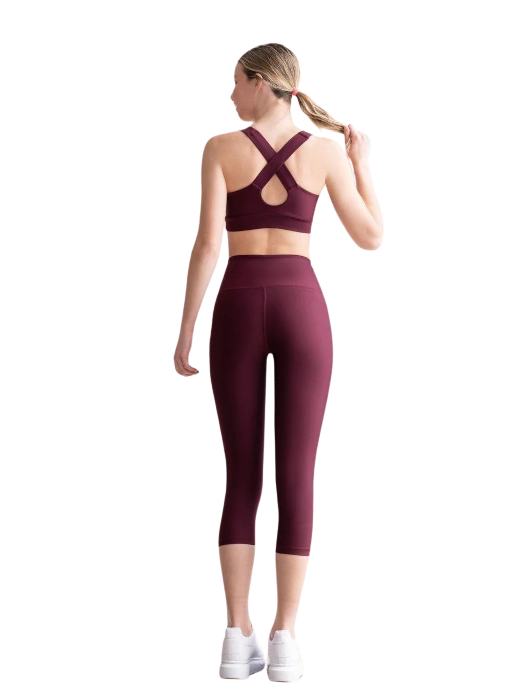 Person in Lismina 4312 Capri-Leggings in Bordeaux und weißen Schuhen, die ihren Pferdeschwanz vor einem weißen Hintergrund zurechtrückt.